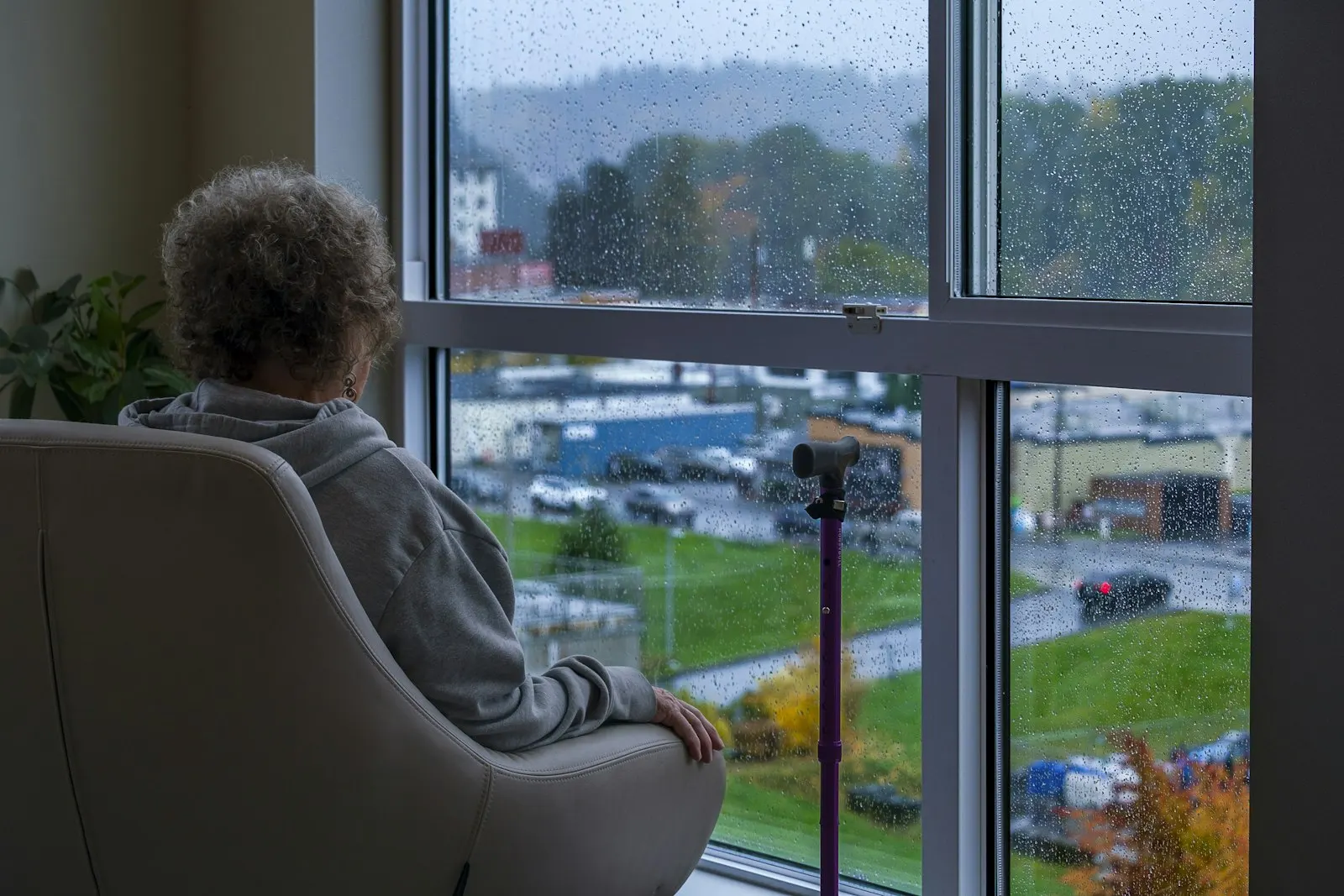 An older woman in an armchair looking out of a rainy window