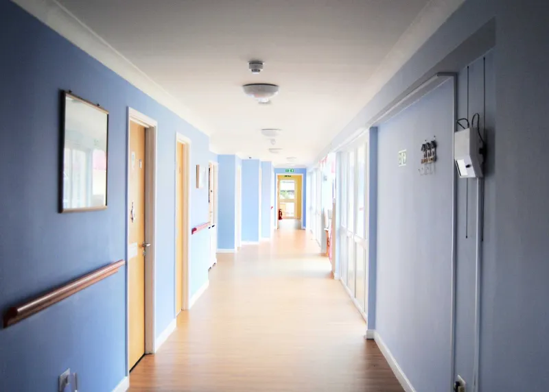 A wide corridor with blue walls, a handrail and wooden doors to residents' rooms
