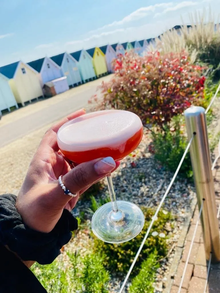 A pink drink held up in front of a row of beach huts and flowers