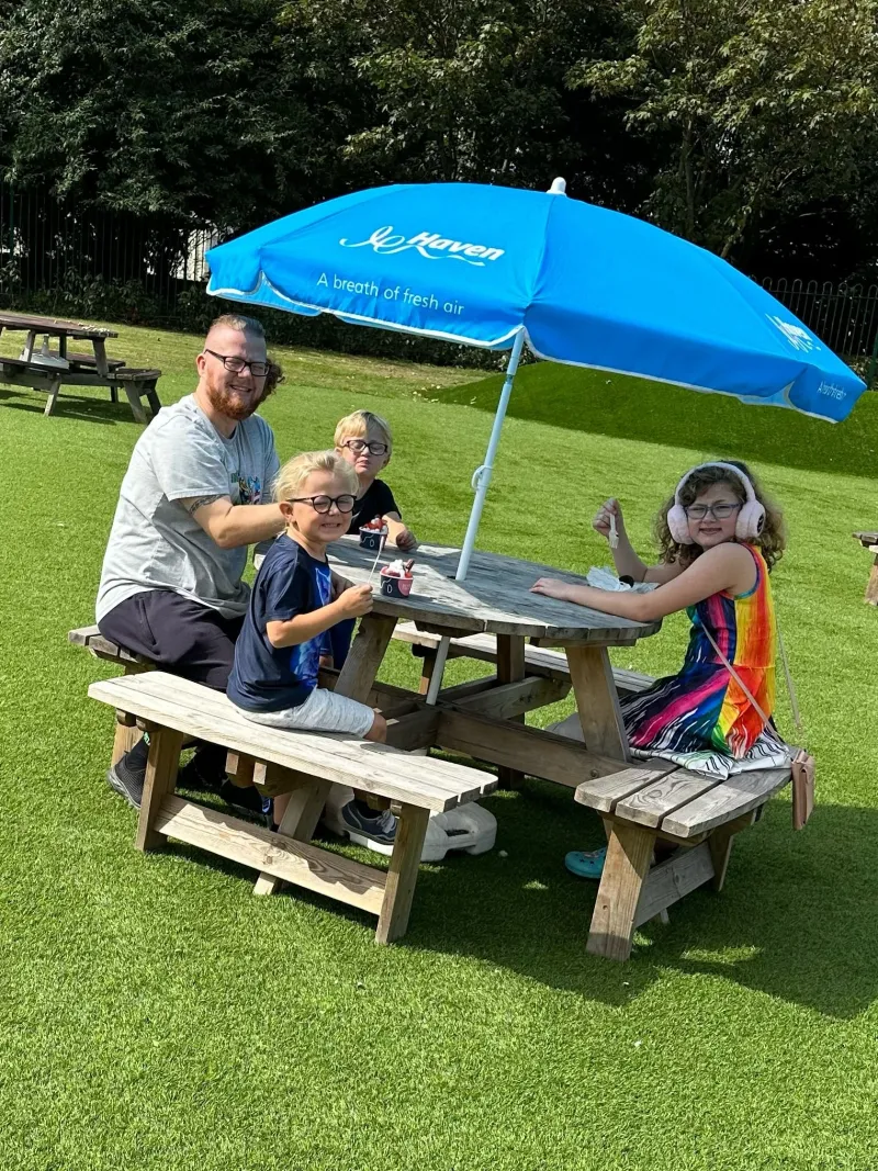 A family at a picnic bench under a blue parasol on the grass