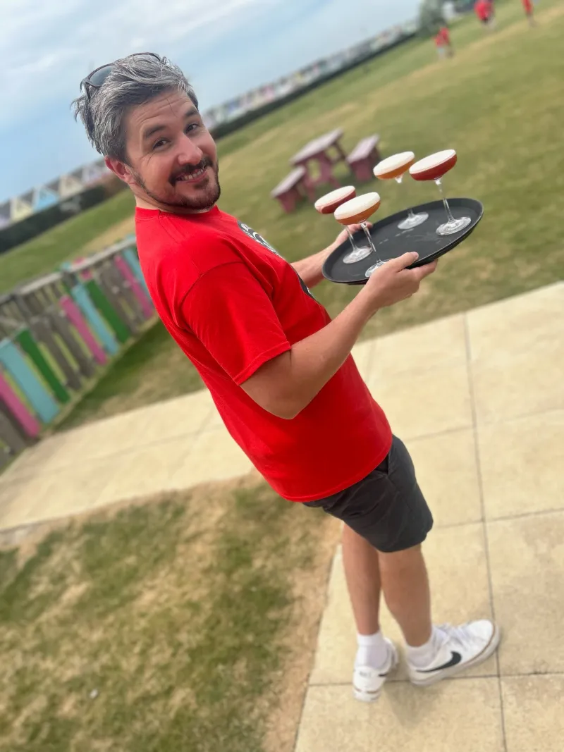 A man in a red T-shirt smiling as he carries a tray of drinks across a lawn