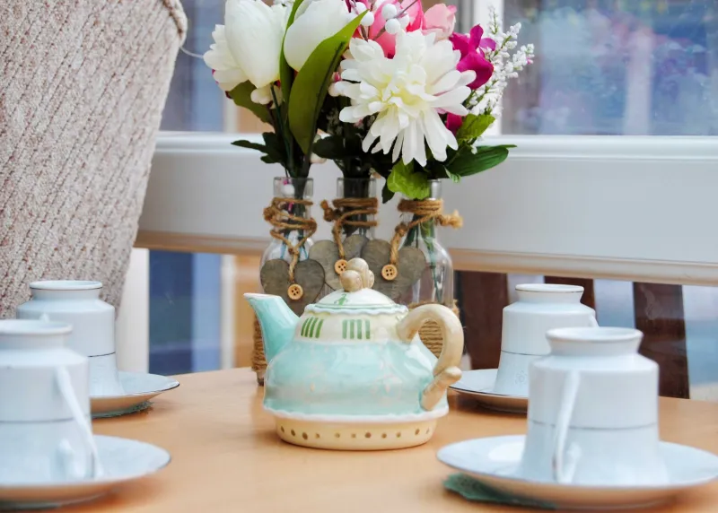 A teapot, cups and saucers and a vase of flowers laid on a table by the window